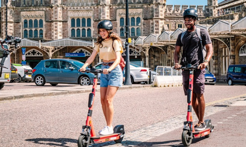 A couple of people on Voi scooters outside Temple Meads, Bristol